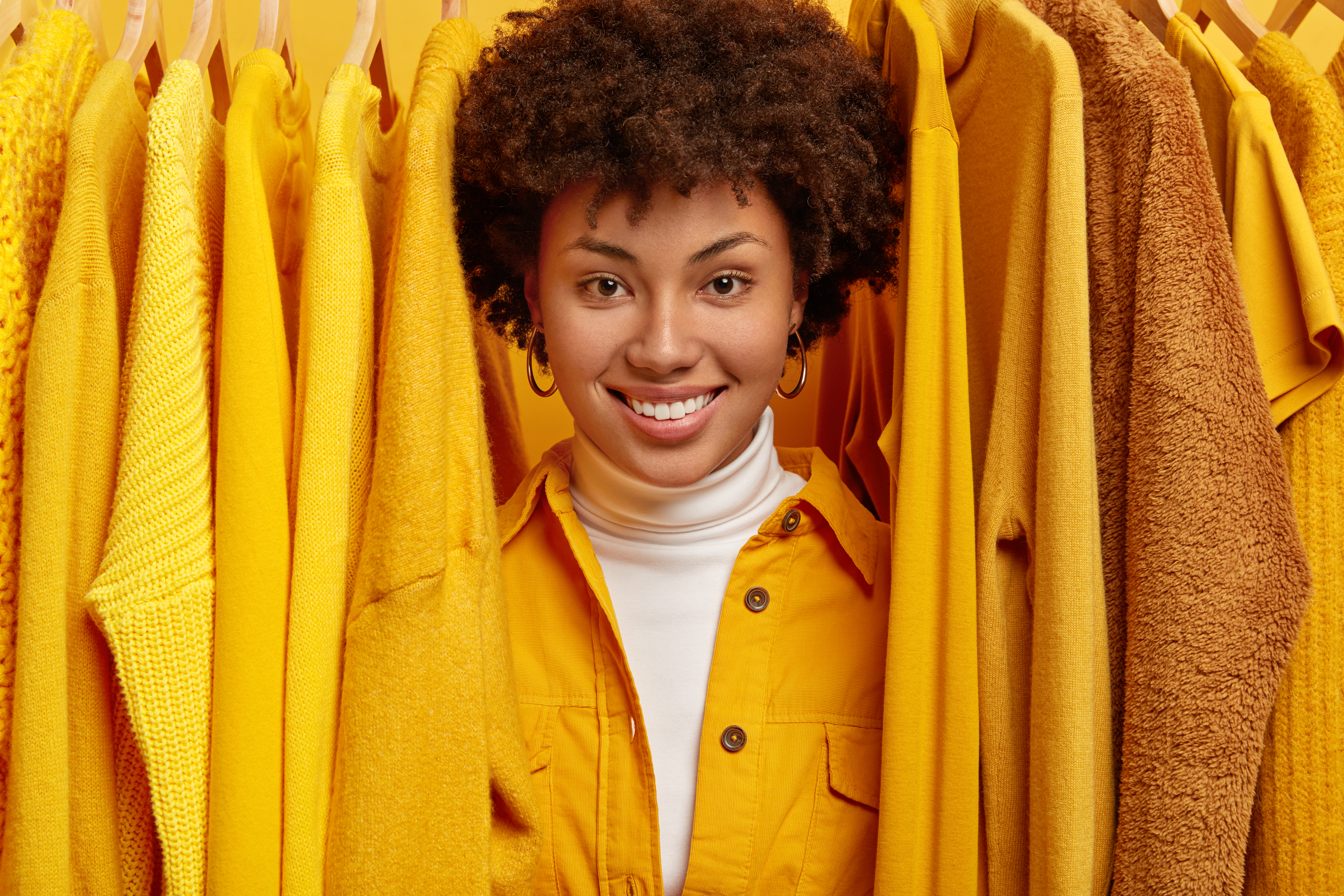 Happy smiling curly haired woman searches what to wear, prefers clothes of yellow color, stands between bright outfits, being real shopaholic, has many outfits in wardrobe, looks through clothing rail